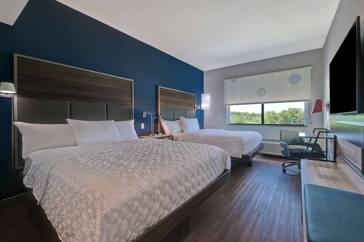 A hotel room with two beds, a desk, a chair, and a large window with a view of greenery.
