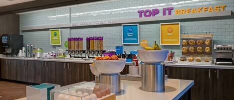 Een ontbijtruimte met een bord waarop 'TOP IT BREAKFAST' staat, en diverse etensopties zoals wafels, eieren en muffins.