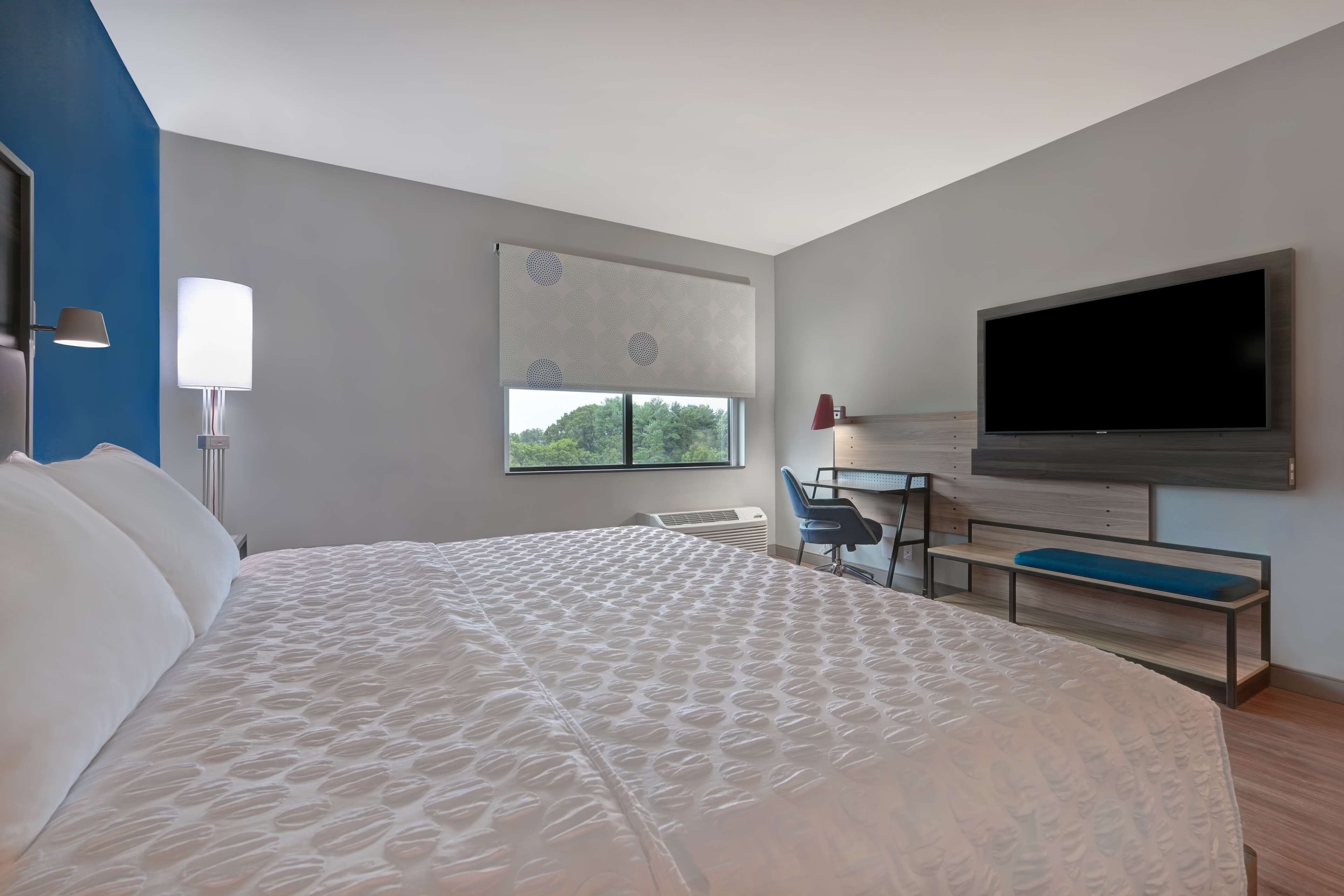 A modern hotel room with a large bed, a desk with a chair, a flat-screen TV, and a window with a view of trees.