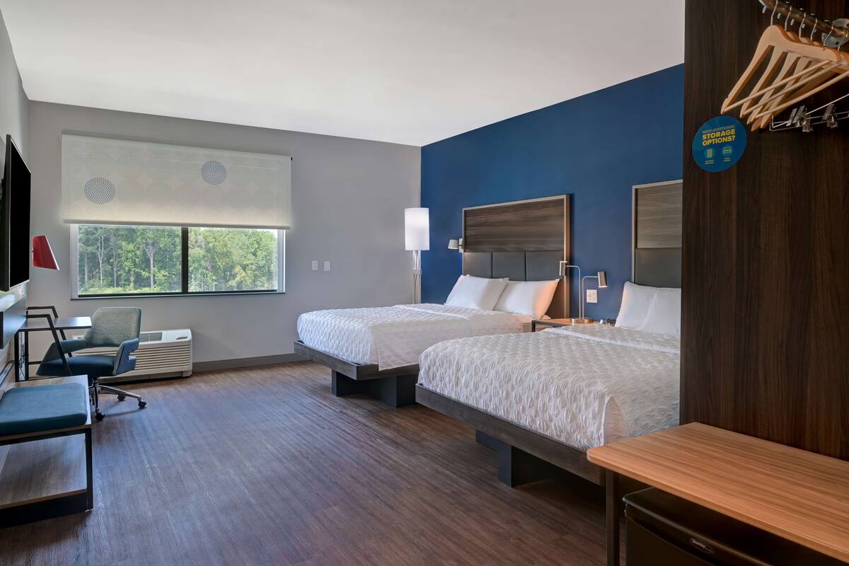 A hotel room with two beds, a desk, a chair, a TV, and a window with a view of trees.