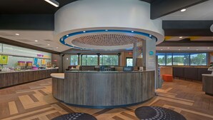 A modern hotel reception area with a curved counter, a view of the outside through large windows, and a seating area with round tables.