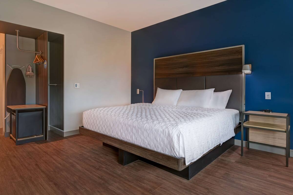 A modern hotel room with a large bed, a bedside table, a wardrobe, and a mirror.