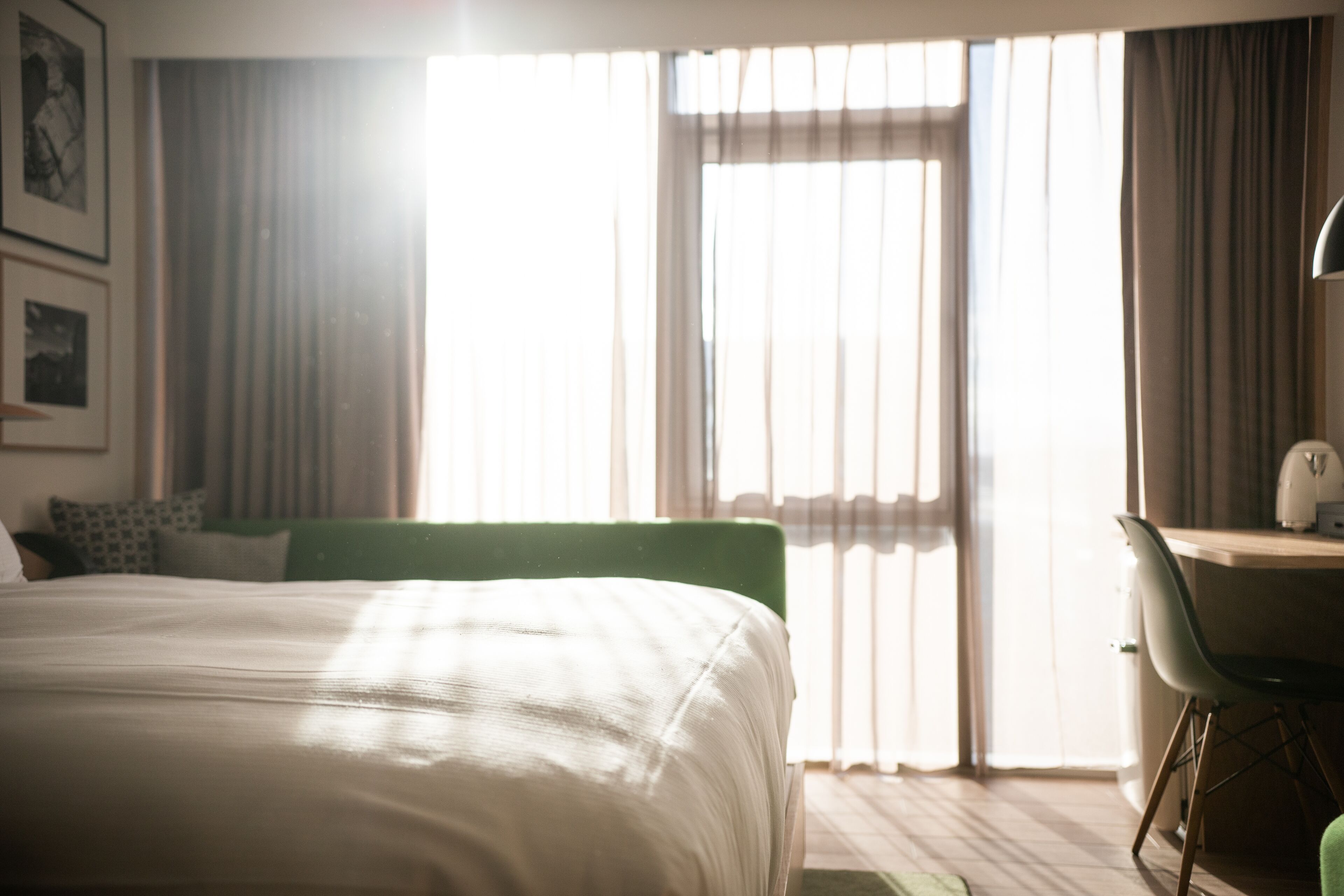 A hotel room with a large bed, a desk, and a chair. There is a window with curtains and a view of the outside.