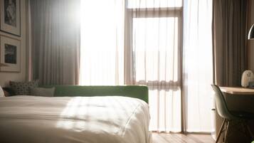 A hotel room with a large bed, a desk, and a chair. There is a window with curtains and a view of the outside.