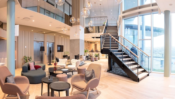 A spacious lobby with a high ceiling, modern furniture, a staircase, and large windows offering views.