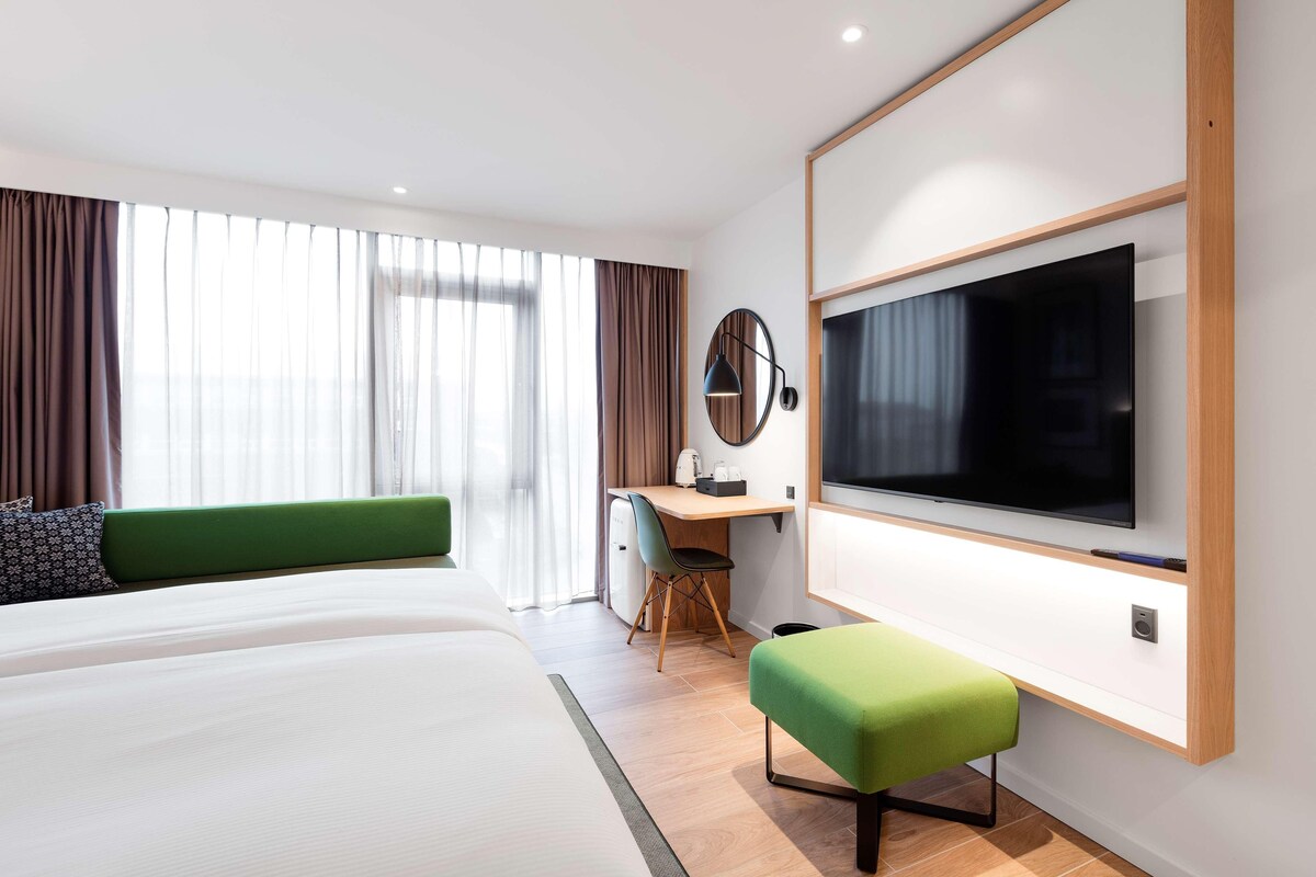 A modern hotel room with a large bed, a desk with a chair, a television mounted on the wall, and a window with curtains.
