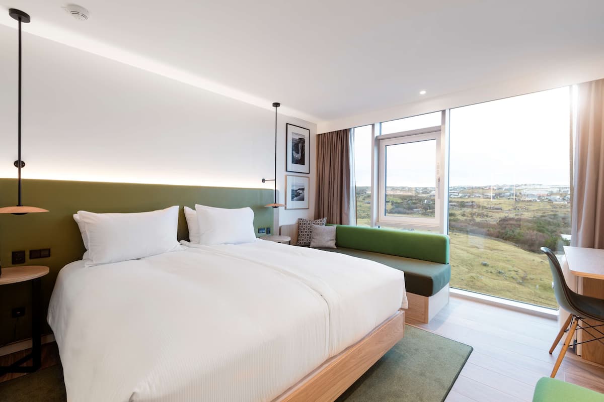 Una habitación de hotel moderna con una cama grande, un sofá verde, una zona de comedor y vistas al campo.