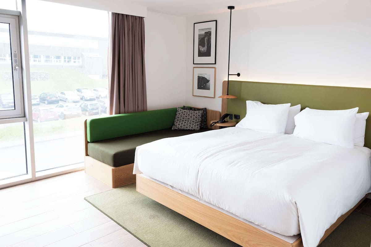 Una habitación de hotel moderna con una cama grande, un sofá verde, un escritorio con lámpara y vistas a coches estacionados a través del ventanal.