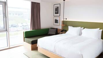 Una habitación de hotel moderna con una cama grande, un sofá verde, un escritorio con lámpara y vistas a coches estacionados a través del ventanal.