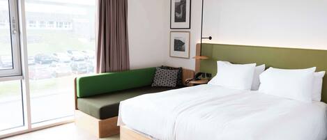 A modern hotel room with a large bed, a green sofa, a desk with a lamp, and a view of parked cars through the window.