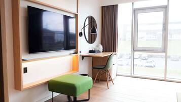 A modern hotel room with a flat-screen TV mounted on the wall, a desk with a chair, a green ottoman, and a large window offering a view of parked cars outside.