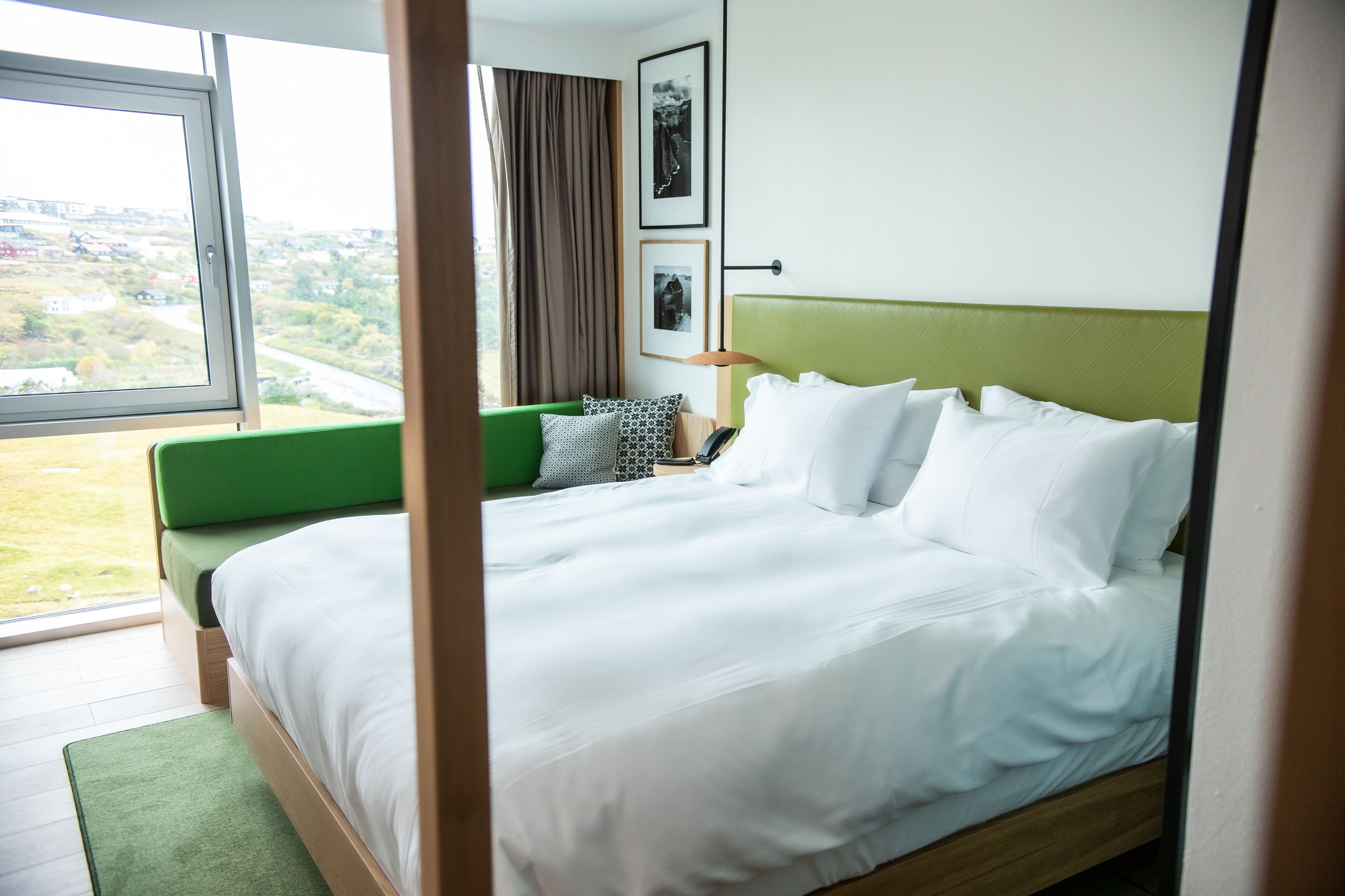 A neatly made bed with white linens, a green headboard, and a view of a cityscape through a window.