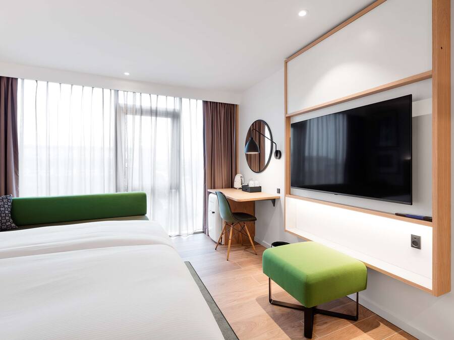 Una habitación de hotel moderna con una cama grande, un escritorio con silla, un televisor de pantalla plana montado en la pared y un ventanal con cortinas.