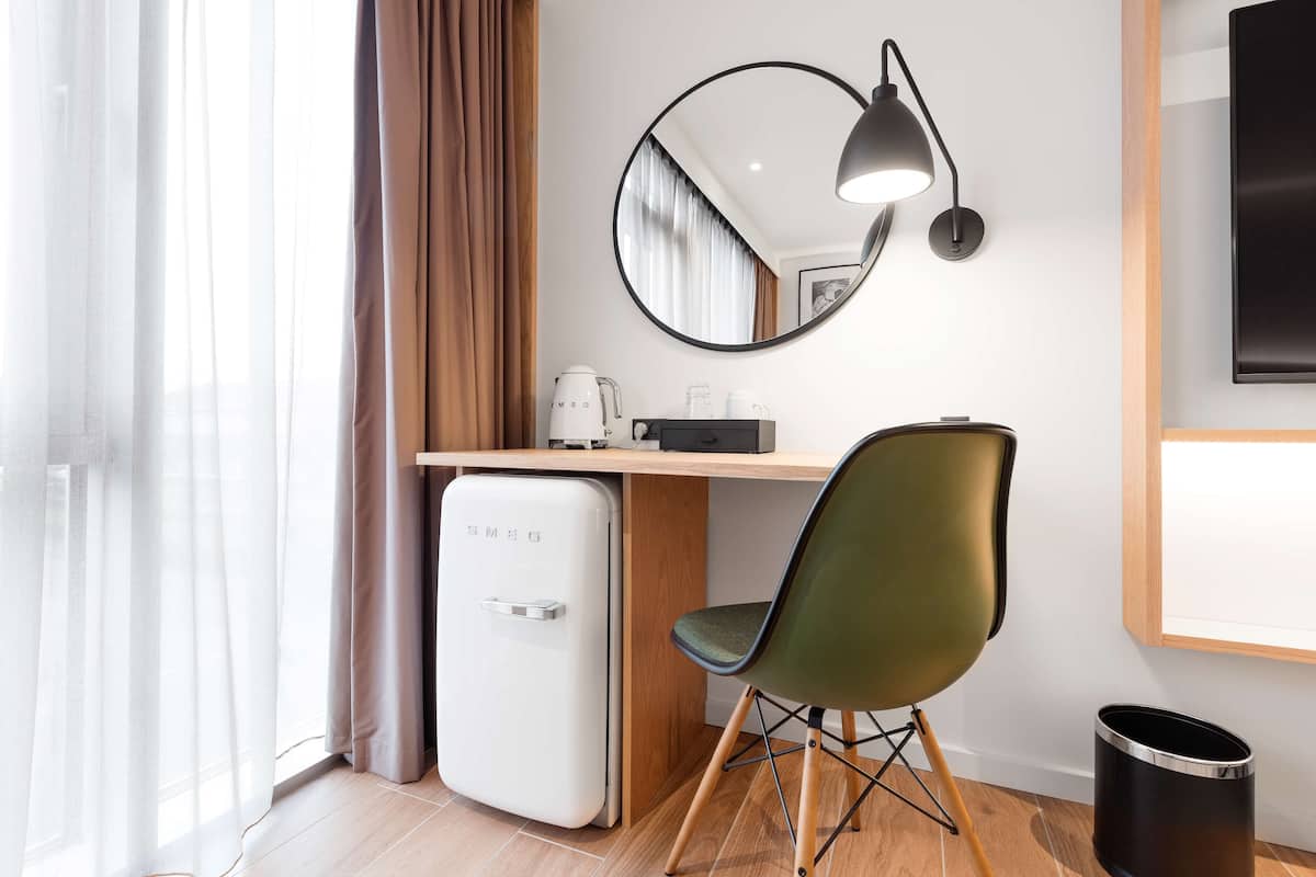 Habitación de hotel compacta con escritorio de madera, refrigerador blanco pequeño, silla verde, basurero negro y televisor instalado en la pared.