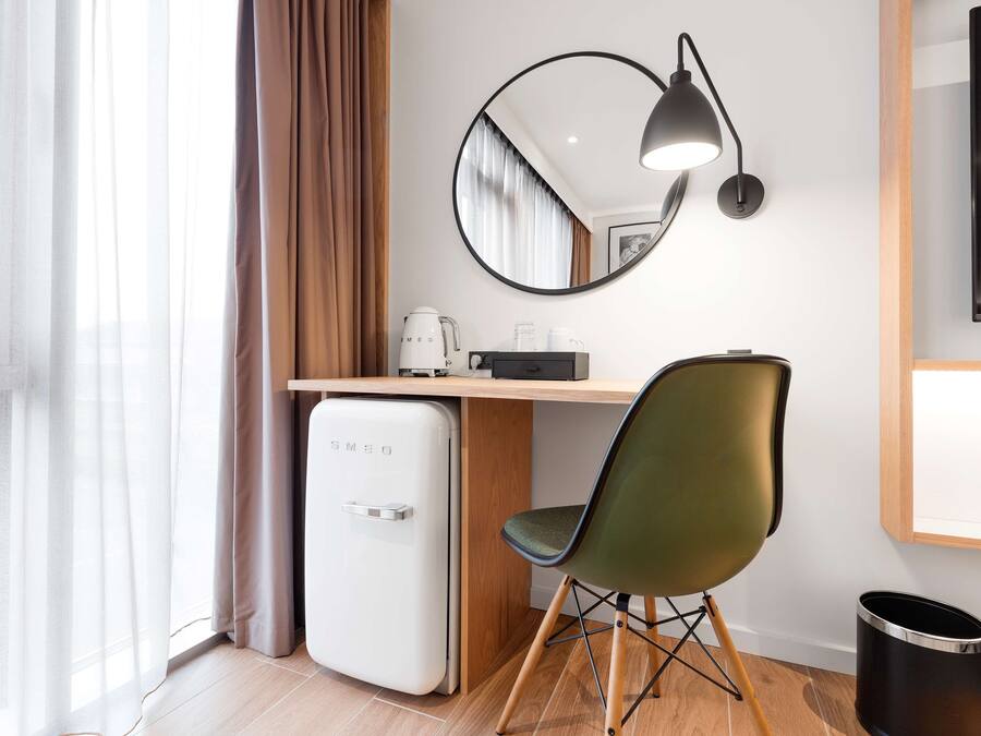 Quarto de hotel compacto com escrivaninha de madeira, frigorífico branco pequeno, cadeira verde, lixeira preta e televisão montada na parede.