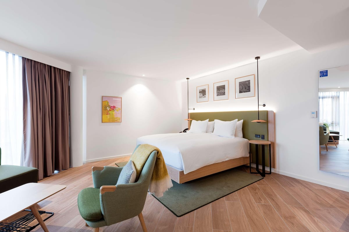 Una habitación de hotel moderna con una cama grande, un sillón verde, una mesita y vistas a la habitación contigua.