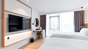 A hotel room with a large flat-screen TV mounted on the wall, a desk with a chair, two beds, and a large window with curtains.