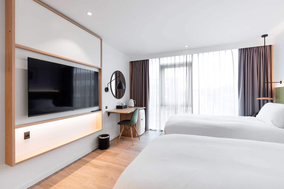 Habitación de hotel con una televisión de pantalla plana grande montada en la pared, un escritorio con una silla, dos camas y un ventanal con cortinas.