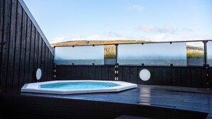 A hot tub on a wooden deck with a view of a hill and a clear sky.