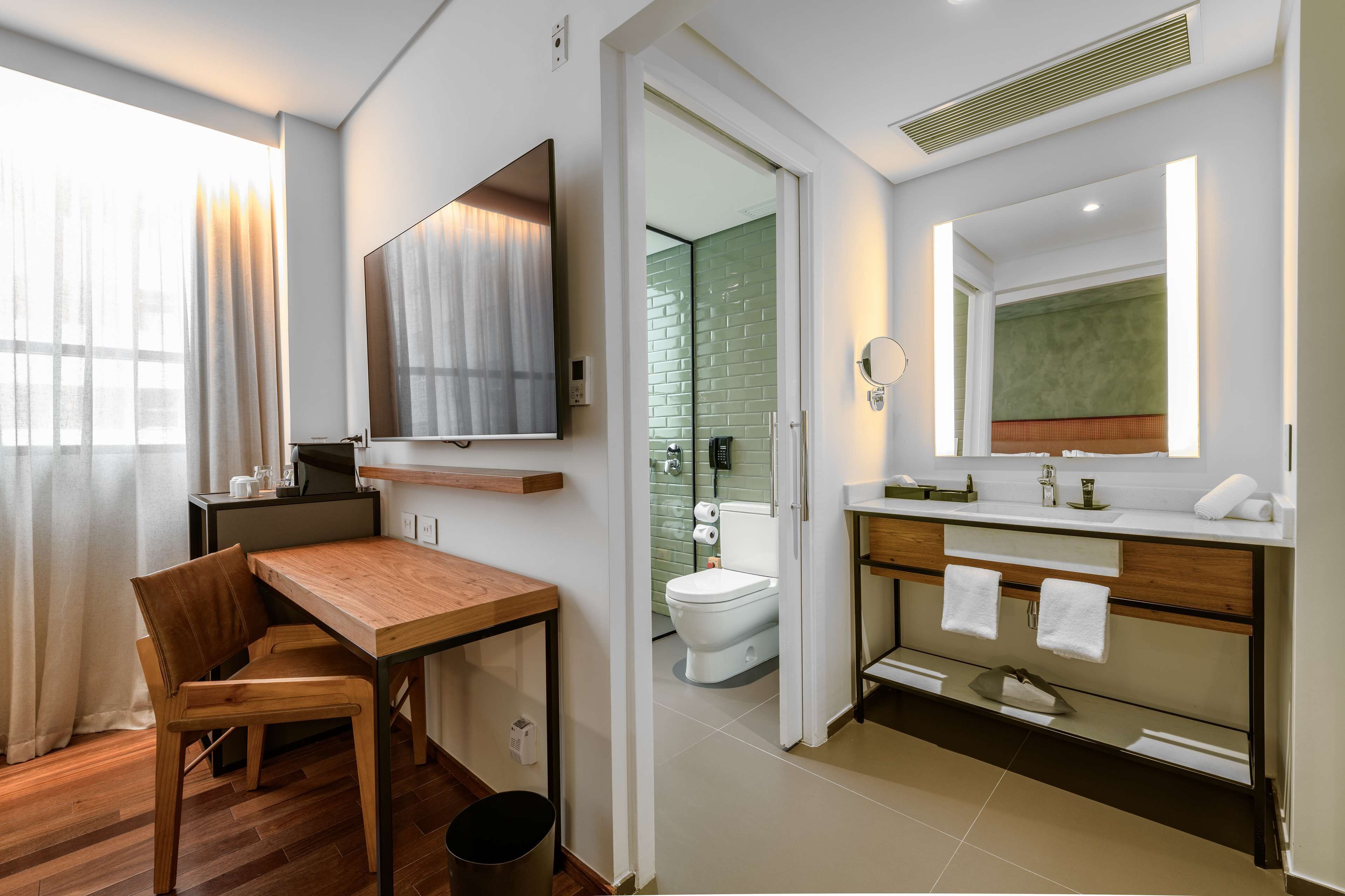 Un baño moderno con inodoro, lavamanos y espejo. En la habitación contigua, hay una mesa de madera y una silla.