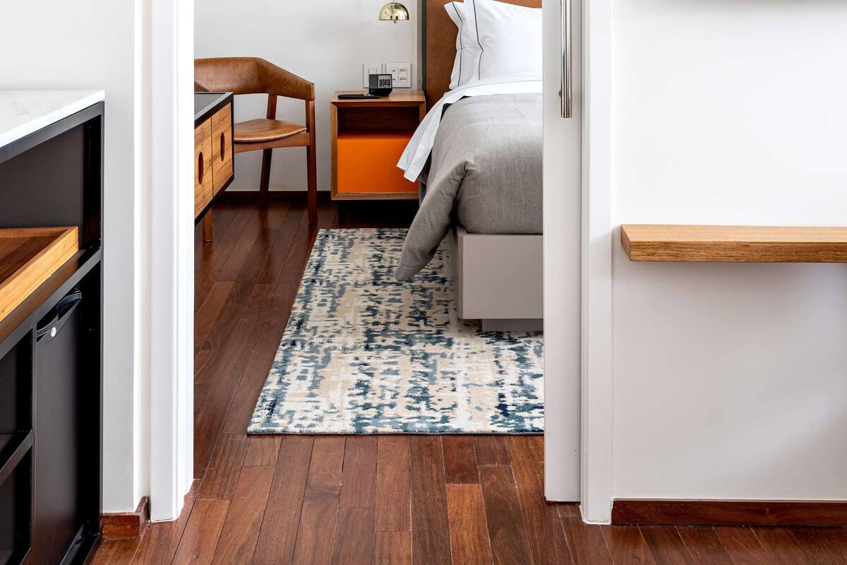 Una habitación de hotel moderna con suelo de madera, una cama con cabecero, un escritorio con silla, una mesita de noche, un televisor y un estante fijado a la pared.