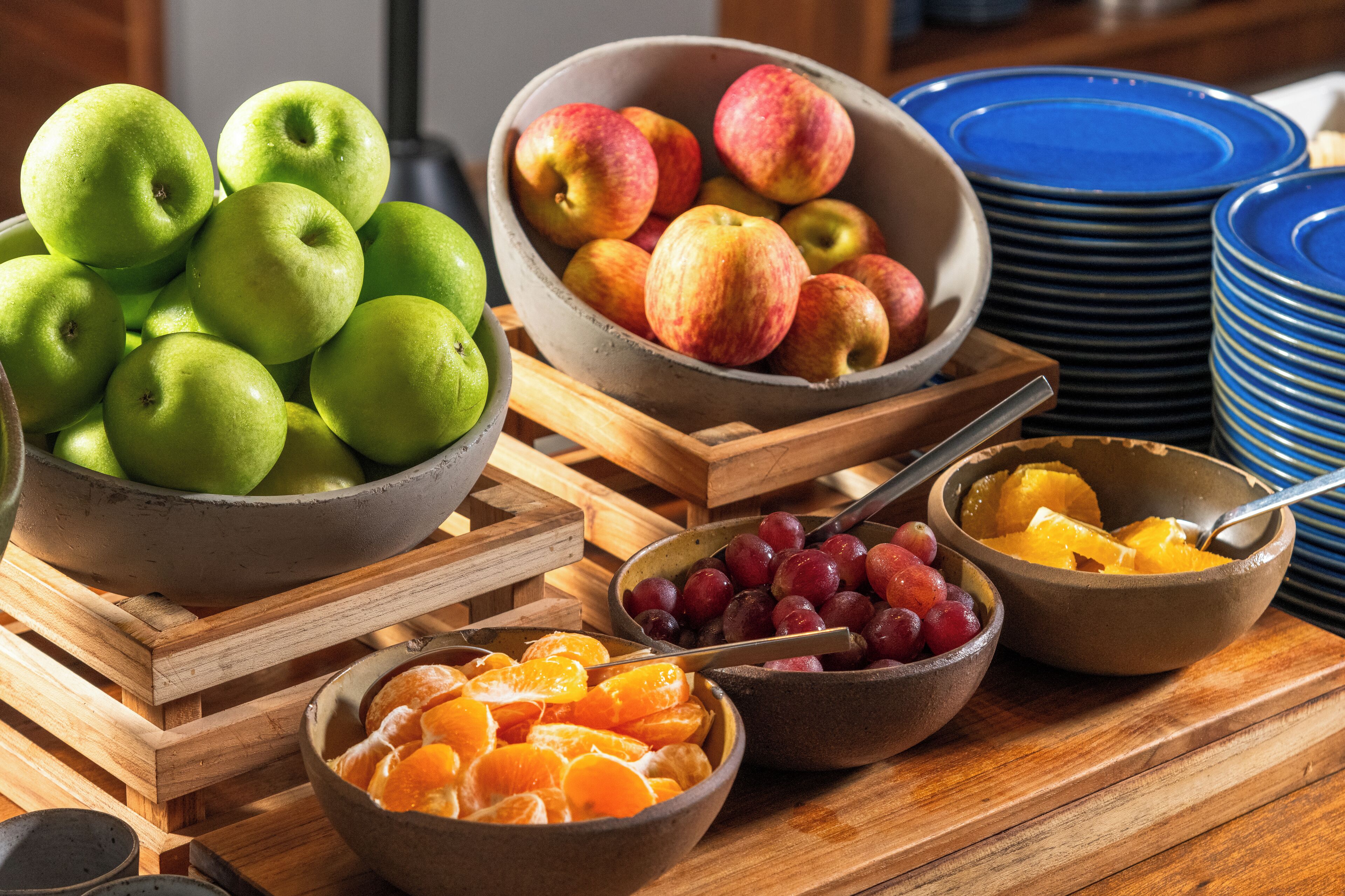 Una mesa de madera con cuencos de manzanas verdes, manzanas rojas, uvas y naranjas.