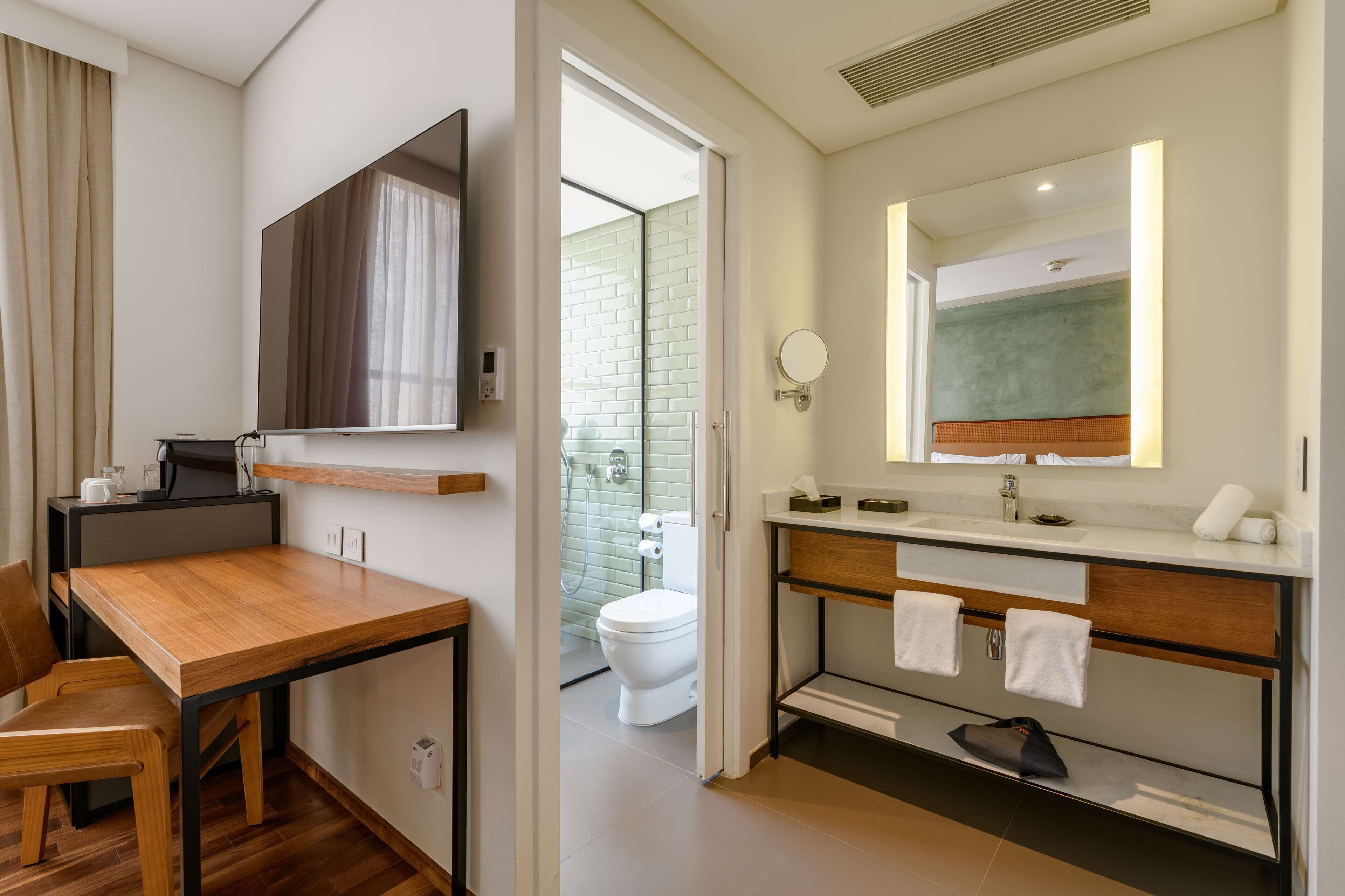 A modern hotel room with a wooden desk, a flat-screen TV, a bathroom with a glass shower, a toilet, and a vanity with a mirror and towel rack.