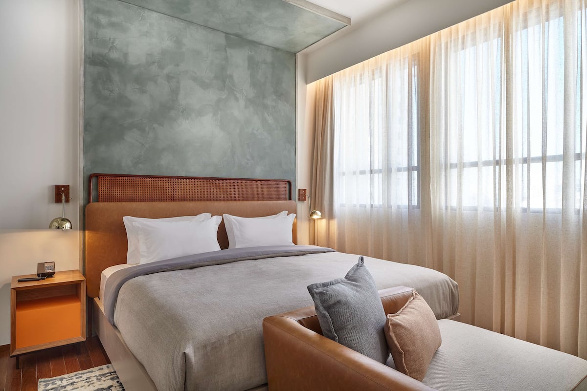Una habitación de hotel moderna con una cama grande, cabecera de madera, mesita de noche con teléfono y cortinas translúcidas en la ventana.