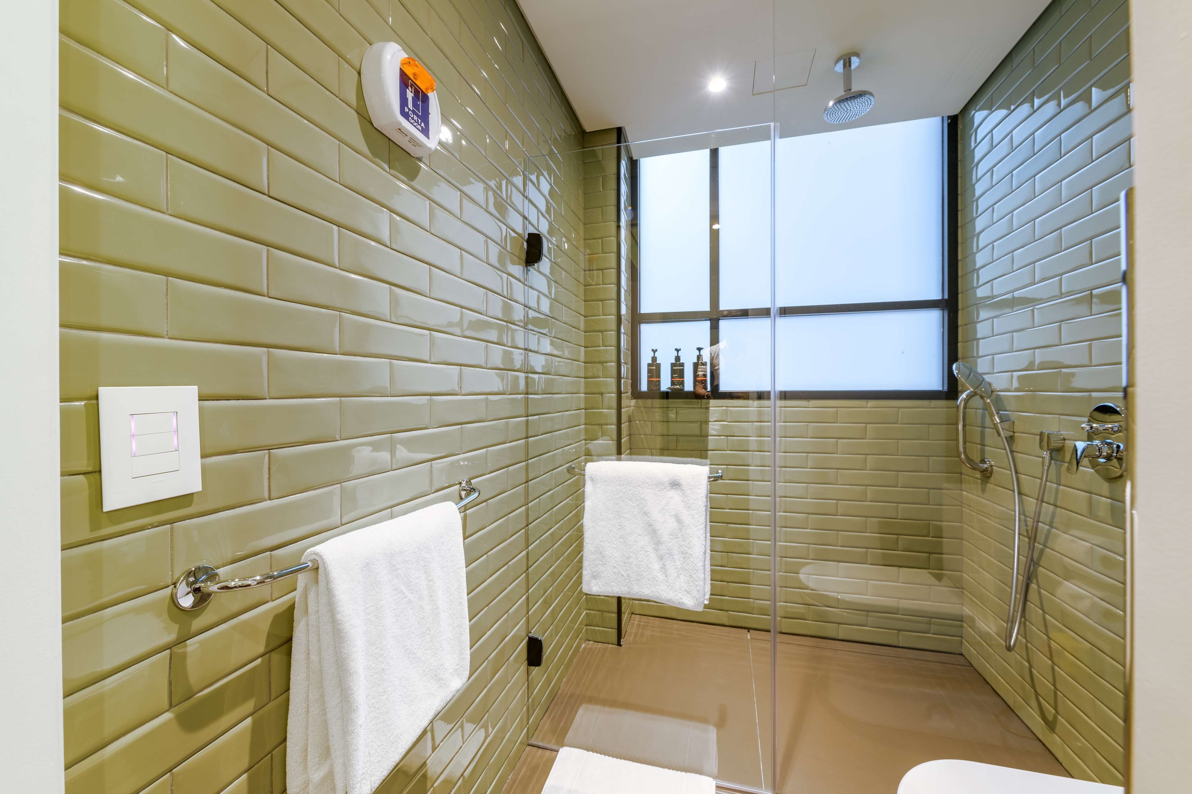A modern bathroom with green tiled walls, a glass shower enclosure, and a window providing natural light.