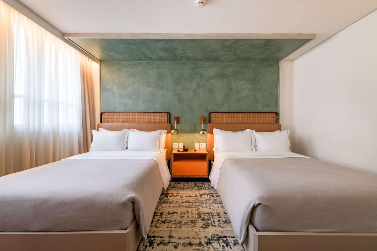 Habitación de hotel con dos camas, una pared de acento verde, un tapete estampado y una ventana con cortinas.