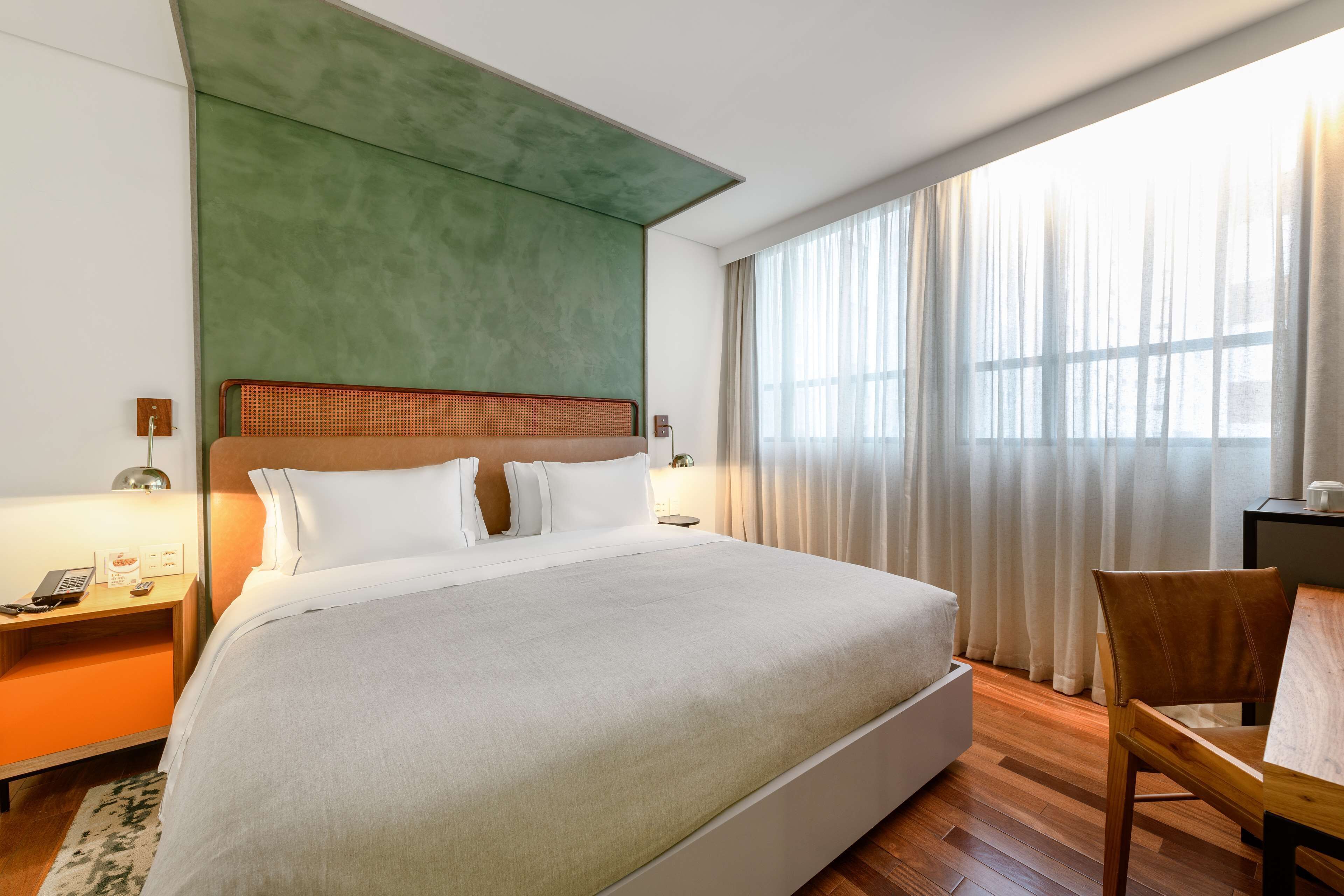 Habitación de hotel con una cama grande, cabecero de madera, un escritorio con silla y un ventanal con cortinas.
