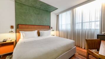 Habitación de hotel con una cama grande, cabecero de madera, un escritorio con silla y un ventanal con cortinas.