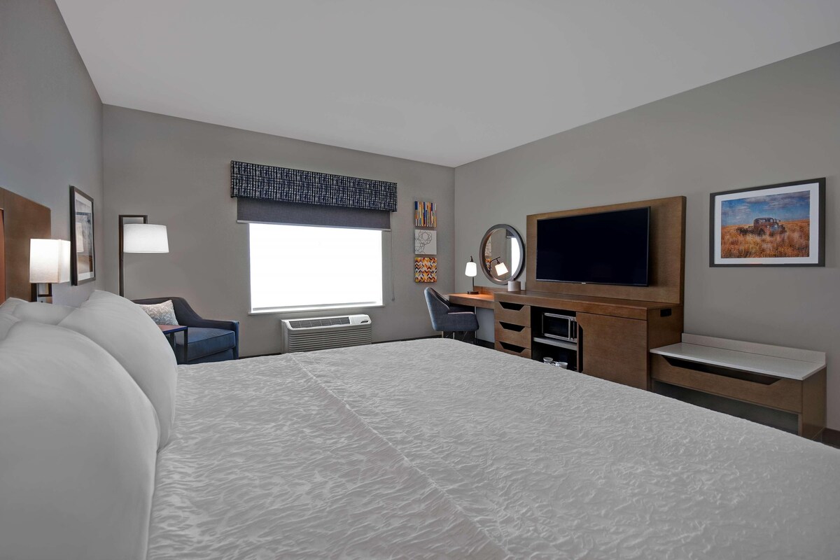 A hotel room with a large bed, a desk with a computer, a TV, and a window with blinds.