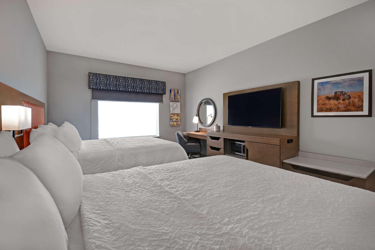 A hotel room with two beds, a desk, a chair, a TV, and a window with a view of a field.