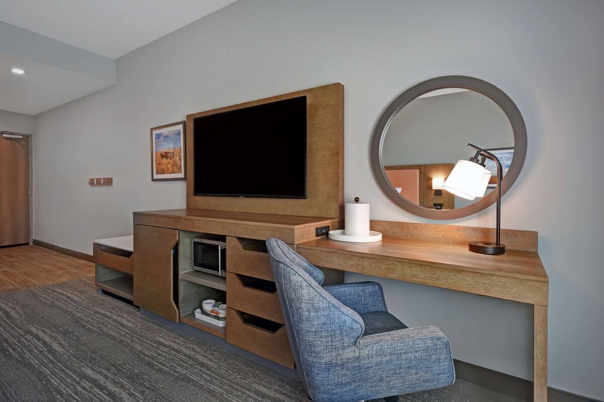 A modern room with a wooden entertainment unit, a flat-screen TV, a round mirror, and a blue upholstered chair.