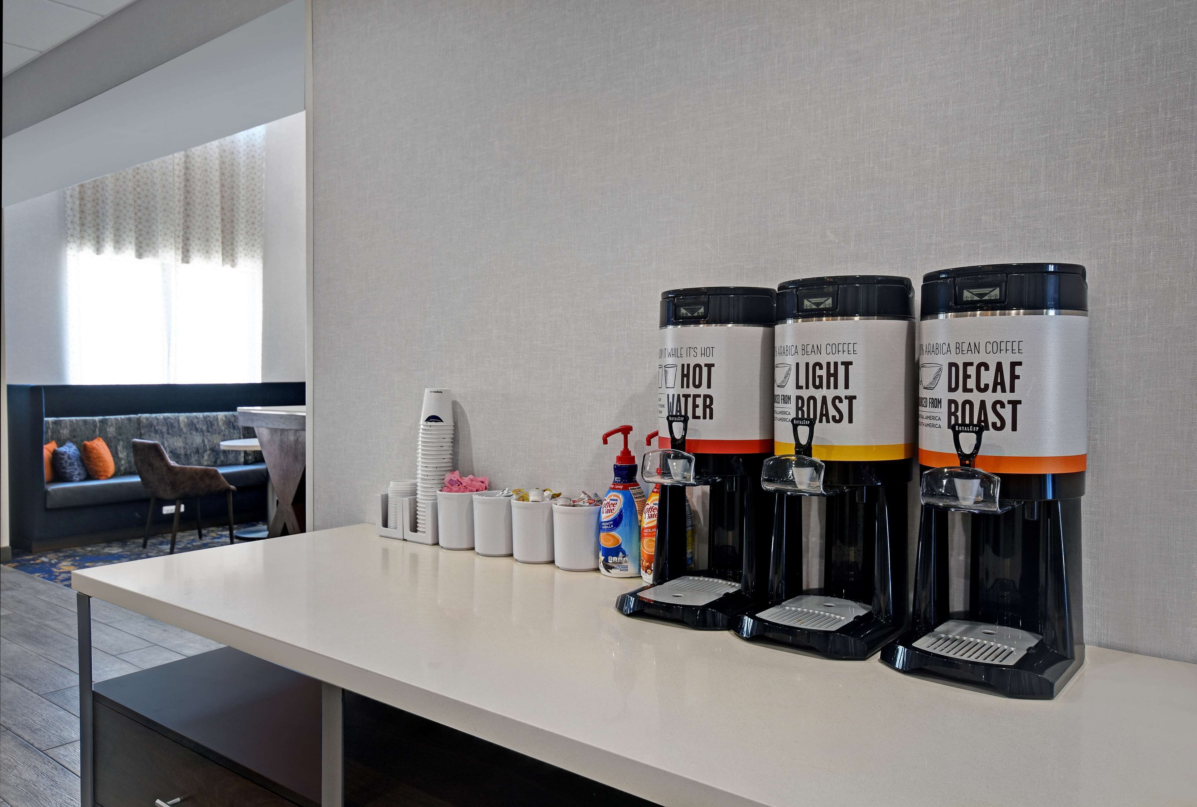 A coffee station with three coffee dispensers labeled 'Hot Water,' 'Light Roast,' and 'Decaf Roast.' There are also cups, a bottle of hand sanitizer, and a tissue box on the counter.