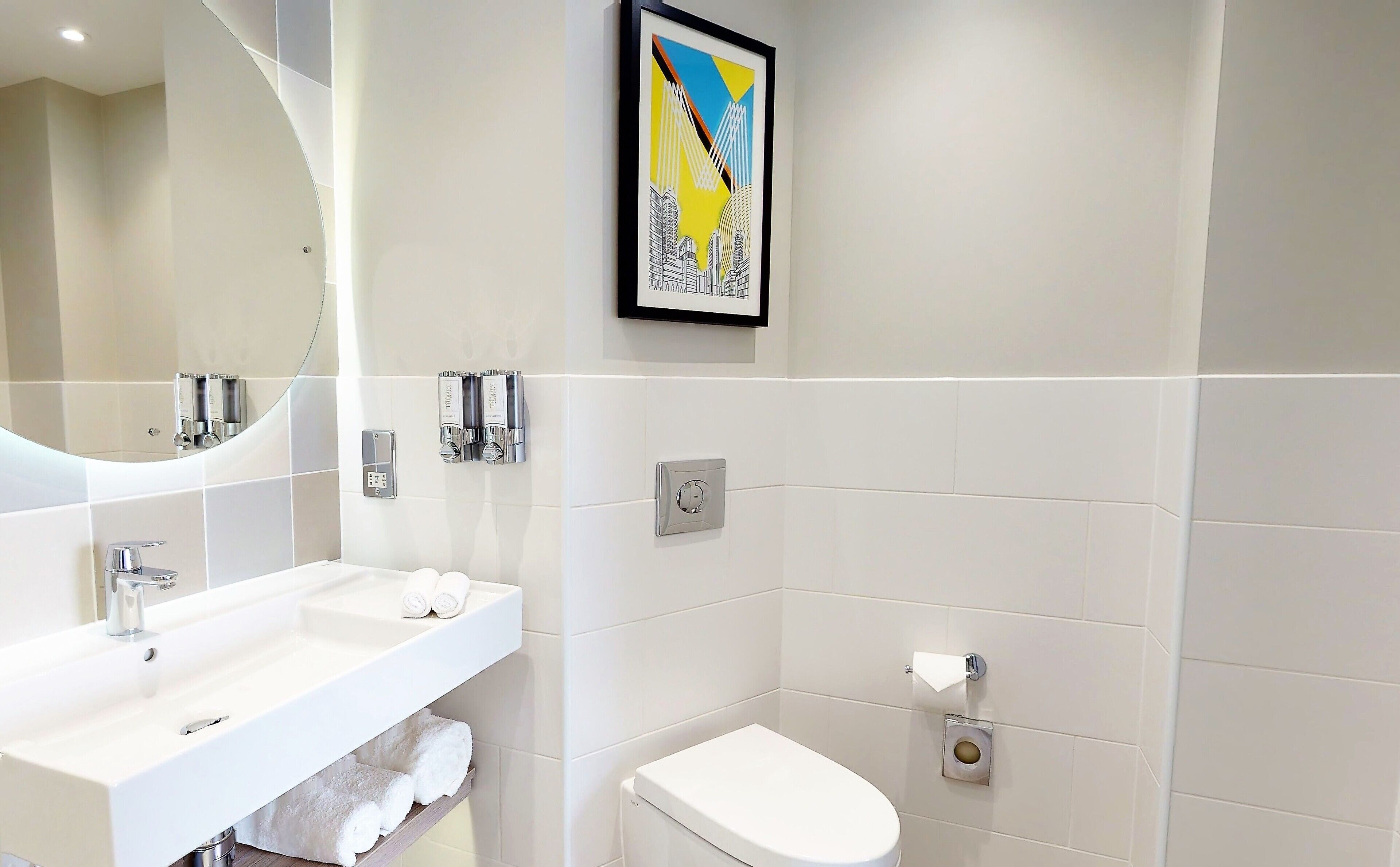 A modern bathroom with a white sink, mirror, and toilet. There is a framed artwork above the toilet and a towel rack with towels.
