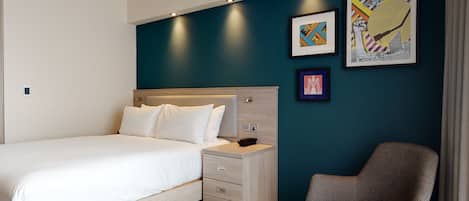 A modern hotel room with a large bed, a nightstand, a chair, and artwork on the wall.