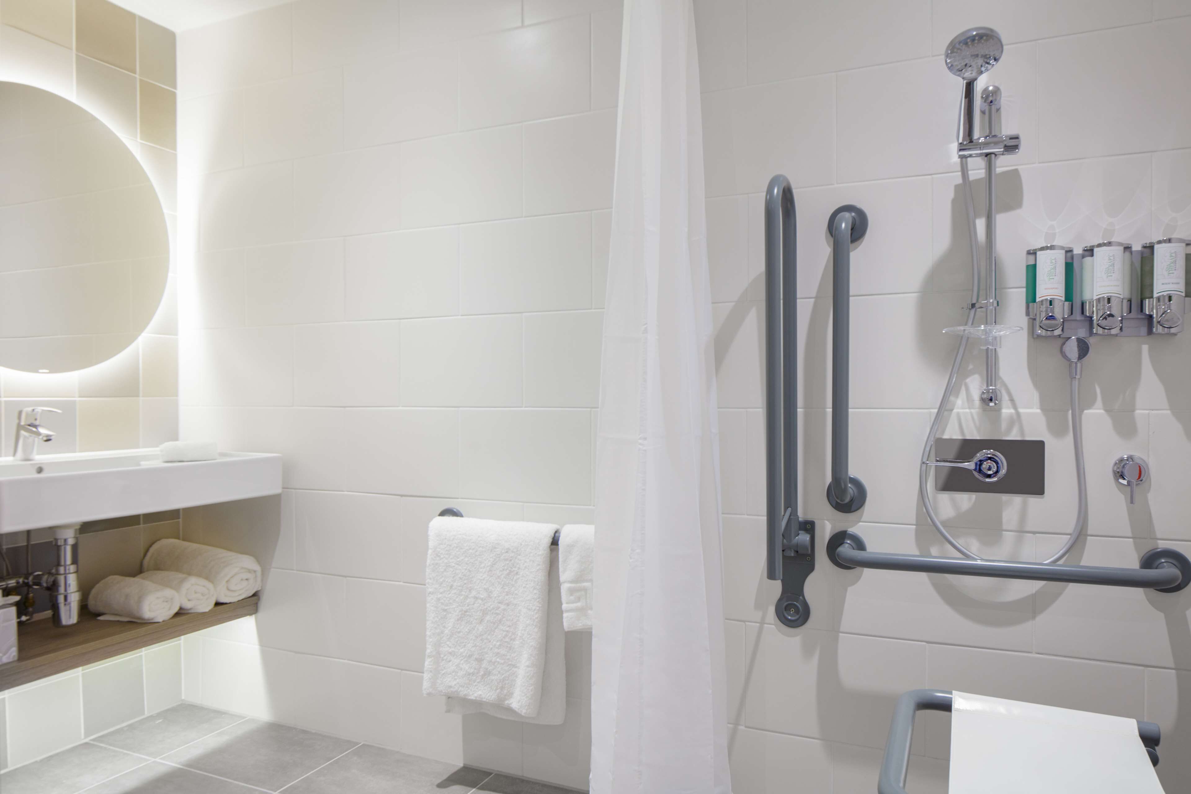 A modern bathroom with a white tiled wall, a sink with a mirror, a towel rack, and a shower with grab bars.