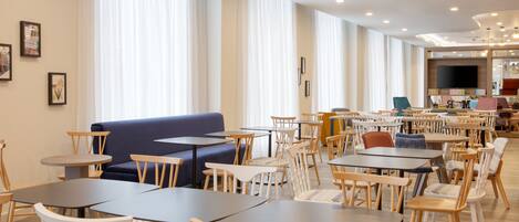 Un vaste espace dédié à la salle à manger, avec plusieurs tables et chaises, un canapé bleu et de grandes fenêtres ornées de rideaux légers.