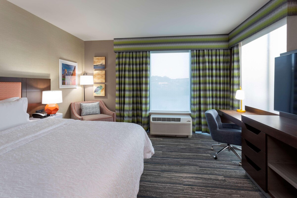 A hotel room with a large bed, a desk, a chair, an air conditioning unit, and a large window with curtains.
