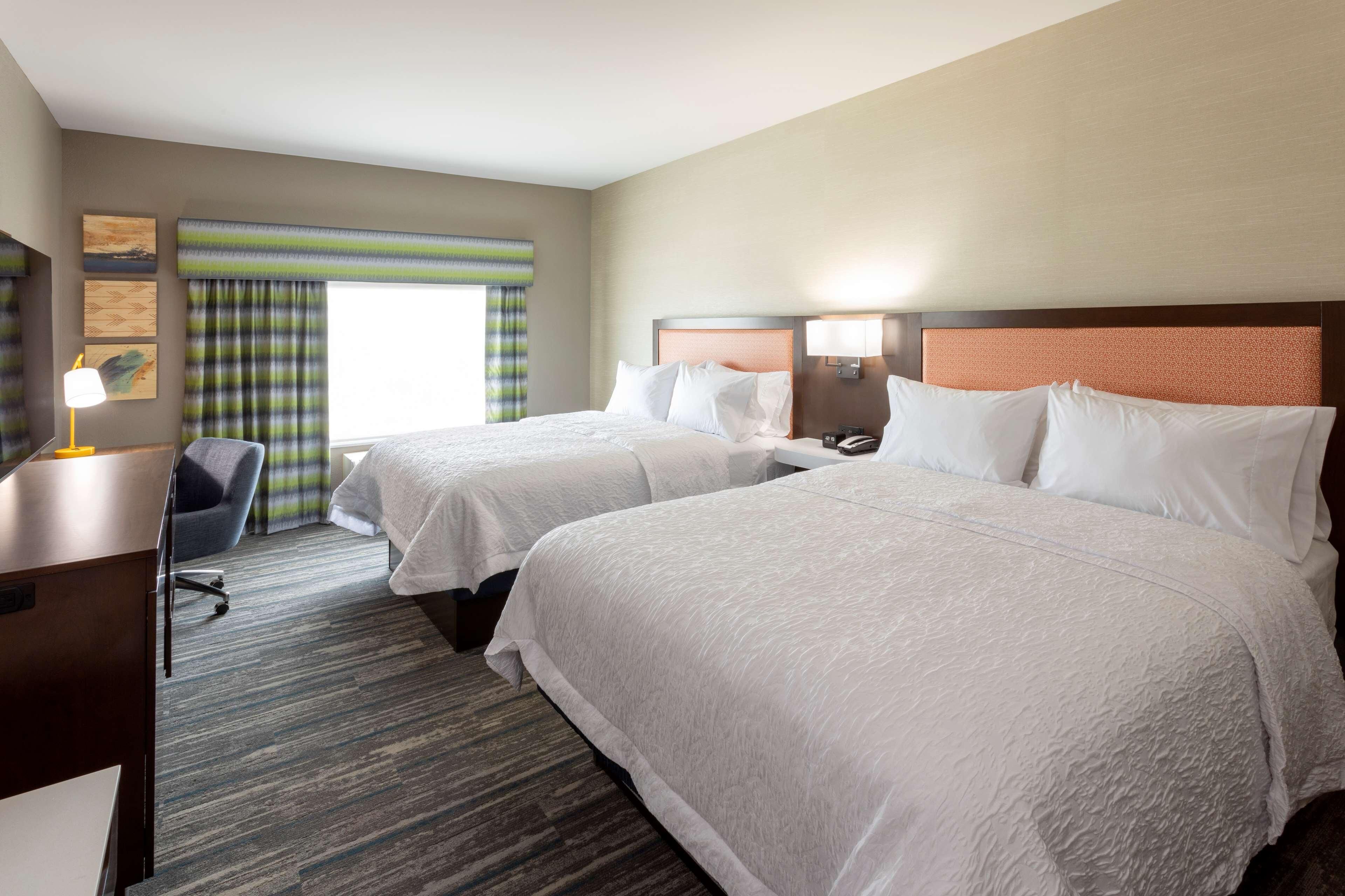 A hotel room with two beds, a desk, a chair, a lamp, and a window with curtains.