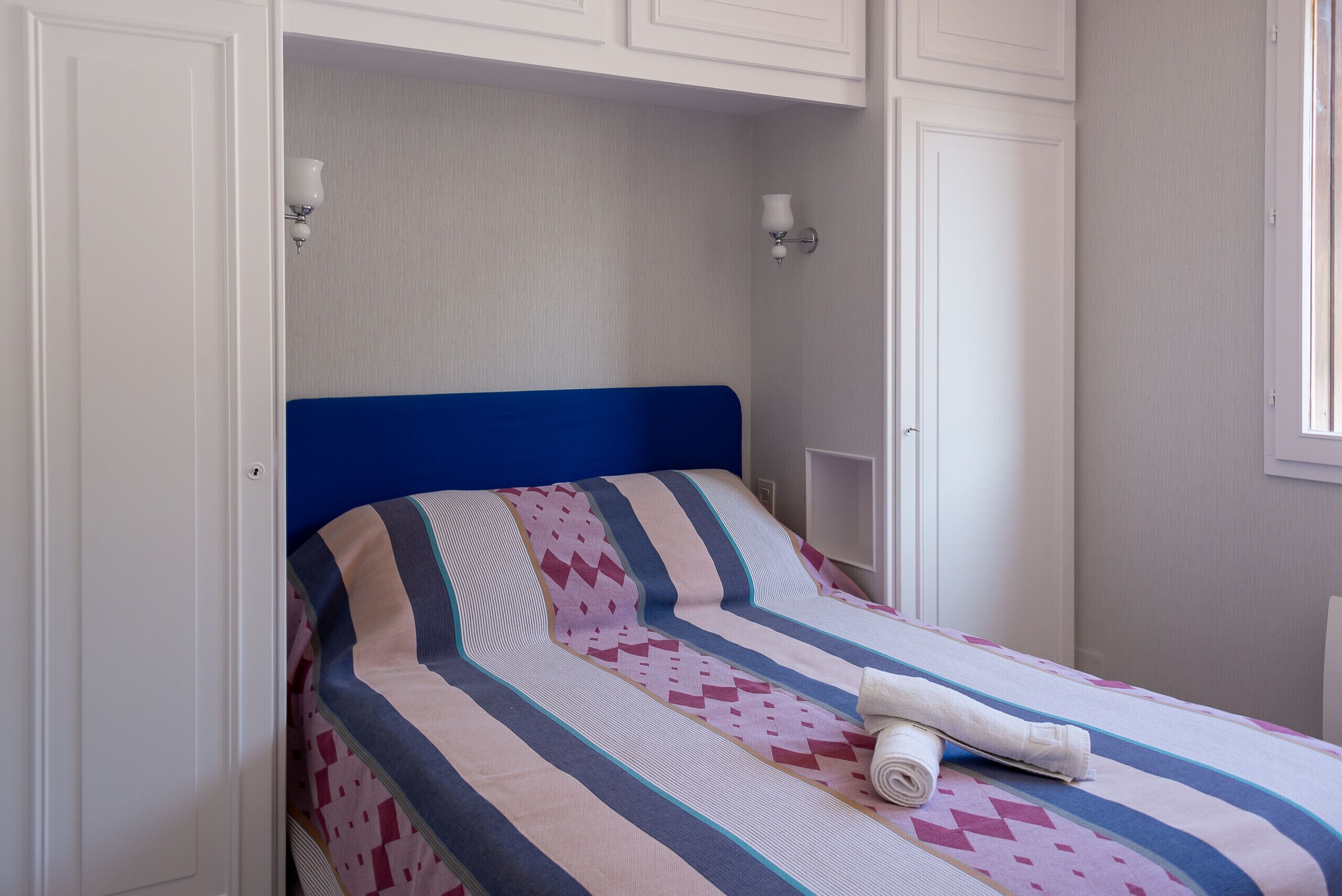 A bed with a blue headboard, striped bedding, and a rolled towel.
