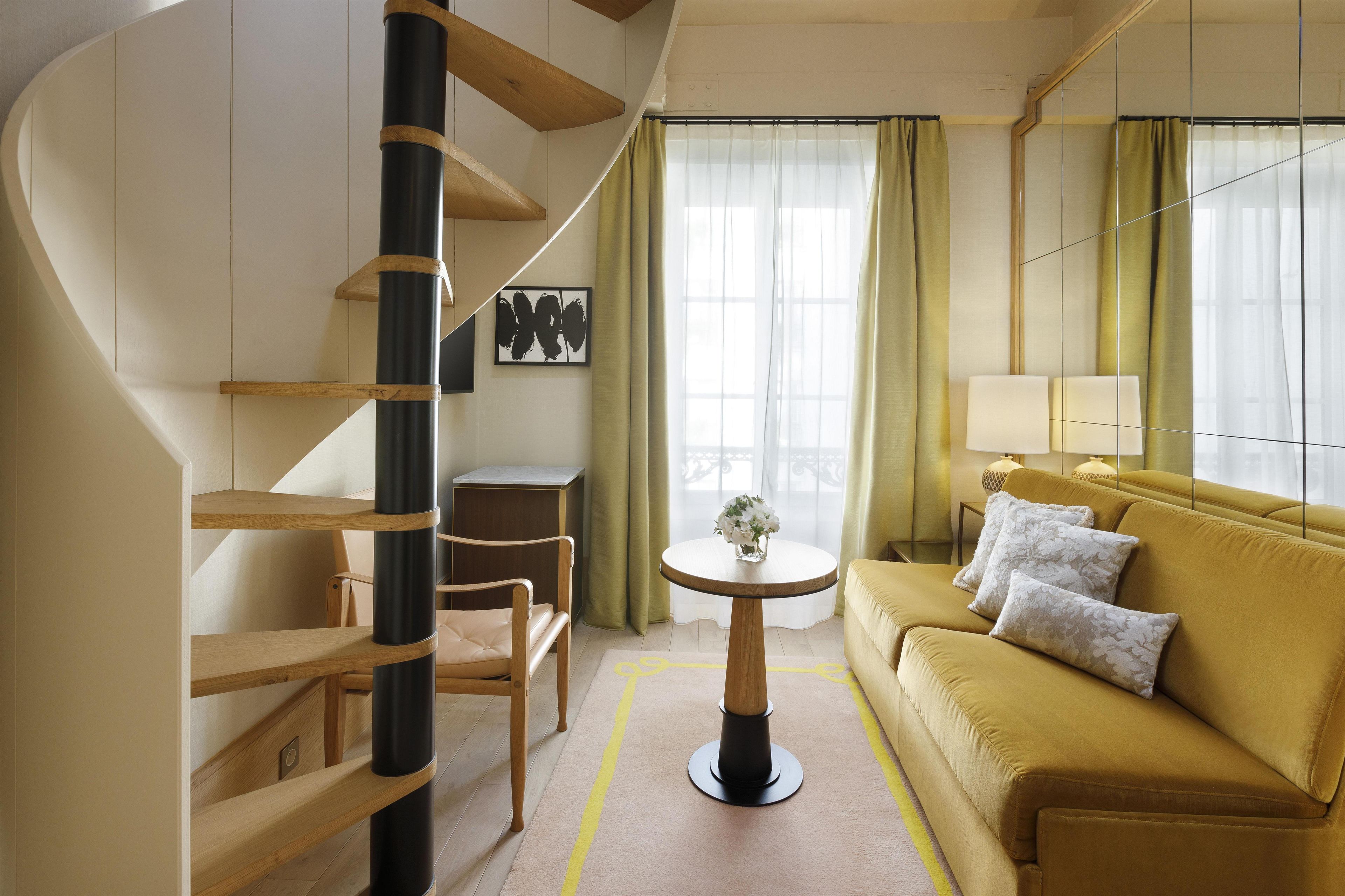 Ett modernt hotellrum med en spiraltrappa, en soffa, ett litet bord med en vas och utsikt över omgivningarna genom stora fönster.