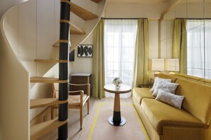 Une chambre d’hôtel moderne avec un escalier en colimaçon, un canapé, une petite table avec un vase, et une vue sur l’extérieur grâce à de grandes fenêtres.