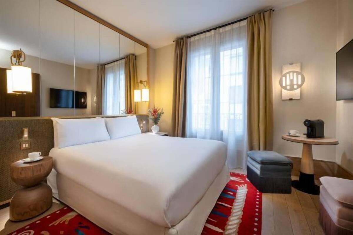 A hotel room with a large bed, a bedside table, a small round table, a chair, a mirror, and a television.