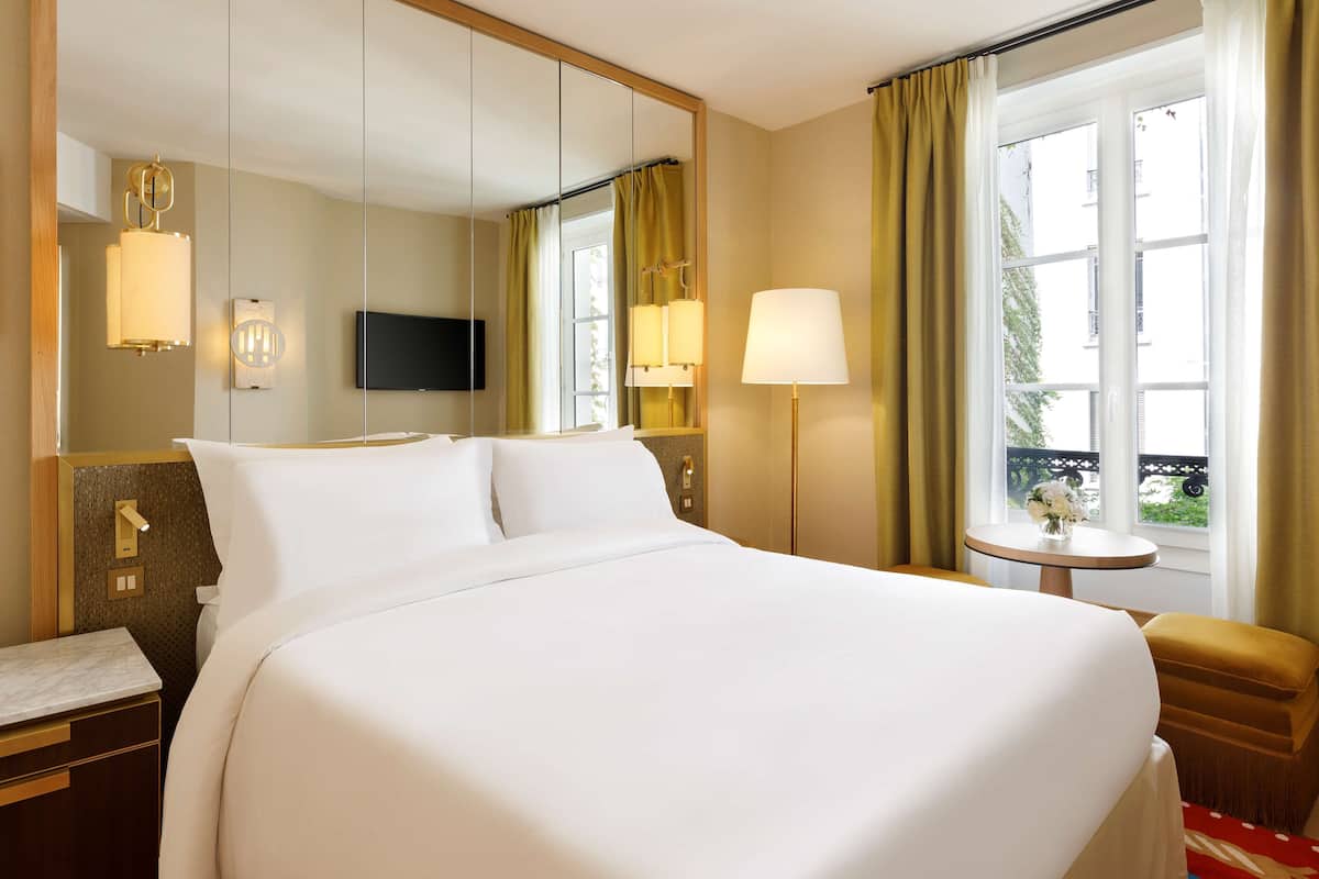 A hotel room with a large bed, a bedside table, a mirror, a TV, and a window with curtains.