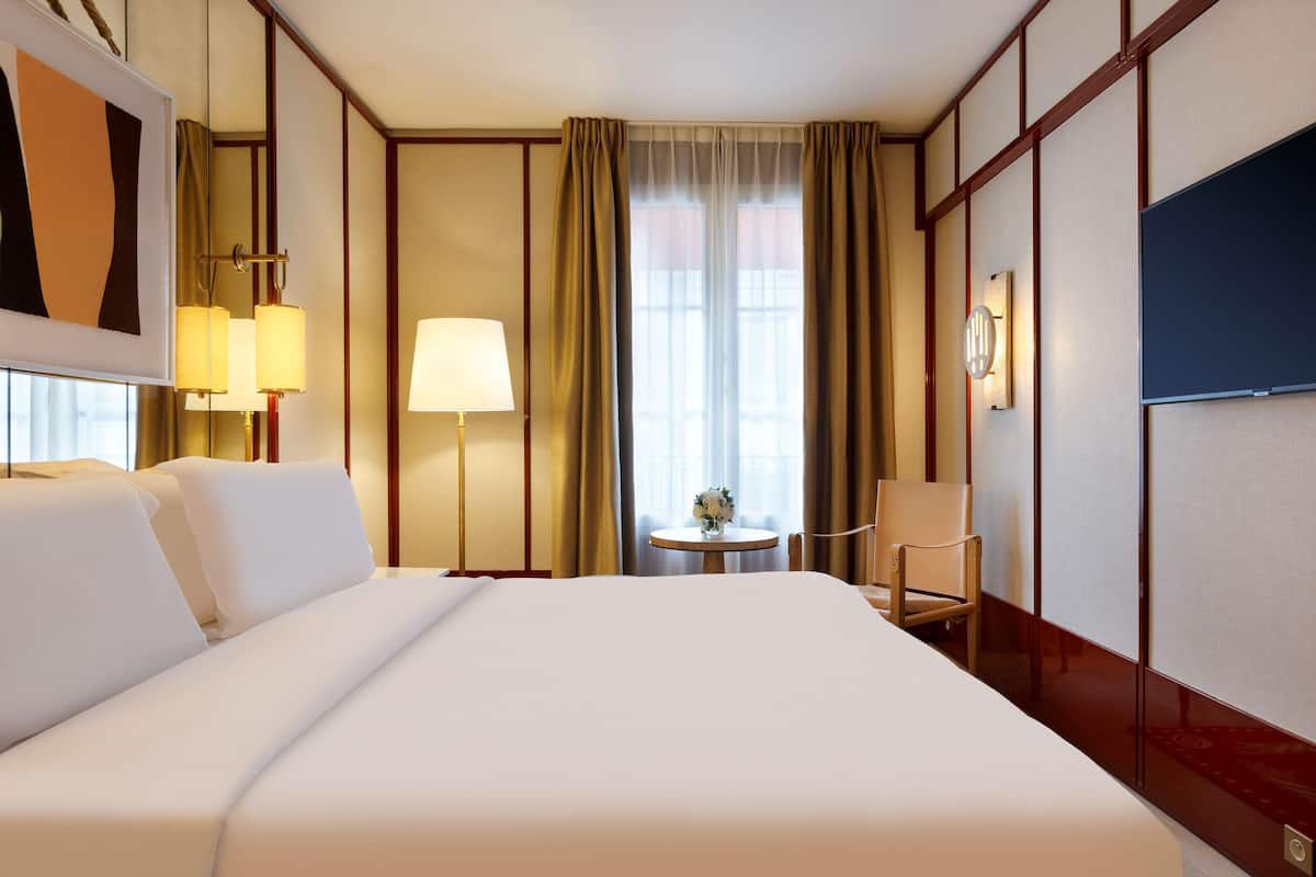 A hotel room with a large bed, a bedside table with lamps, a chair, a small round table with a vase, and a large painting on the wall.