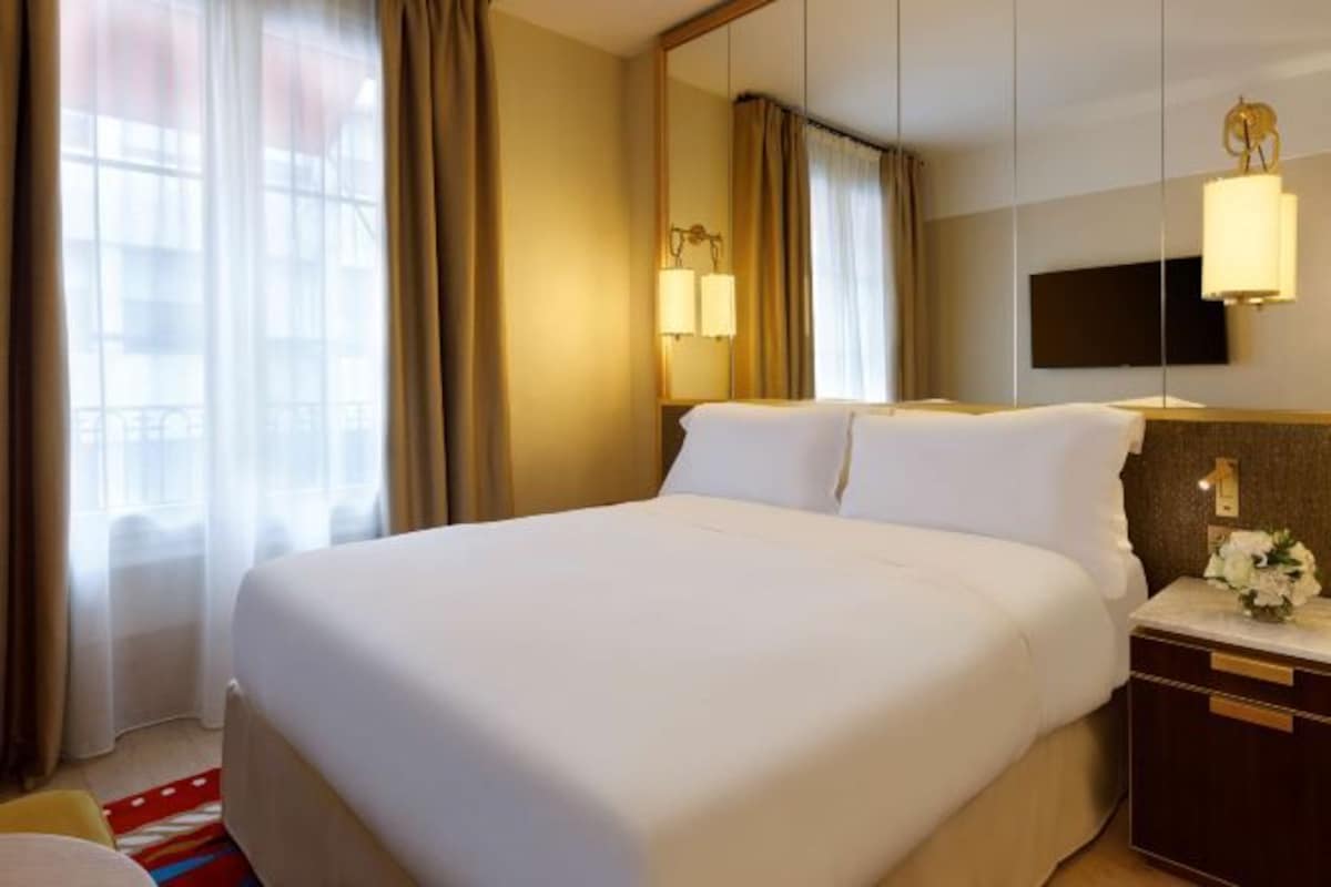 Ein Hotelzimmer mit einem großen Bett, einem Nachttisch mit Lampe, einem an der Wand befestigten Fernseher und einem Fenster mit Vorhängen.