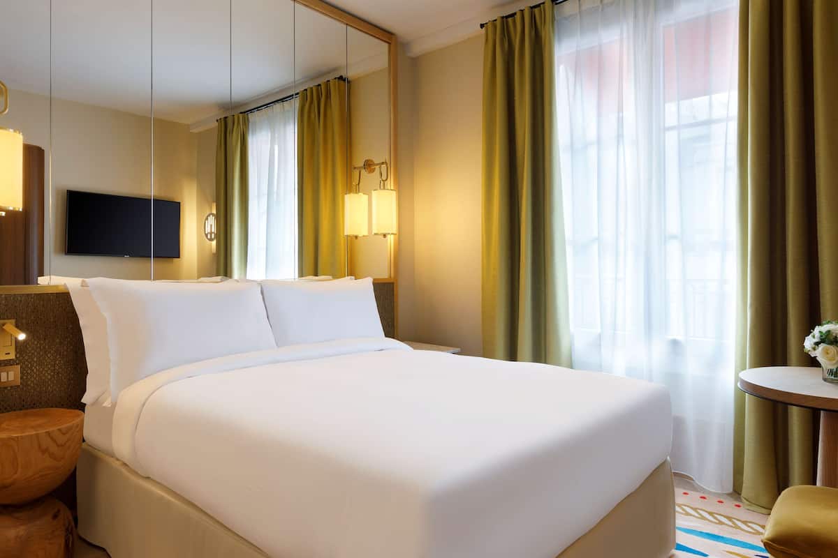 A hotel room with a large bed, a flat-screen TV mounted on the wall, a bedside table with a lamp, and a small round table with a vase of flowers.